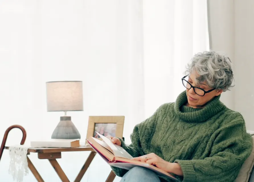 Eine Seniorin liest in einem hellen Wohnzimmer in einem Buch.